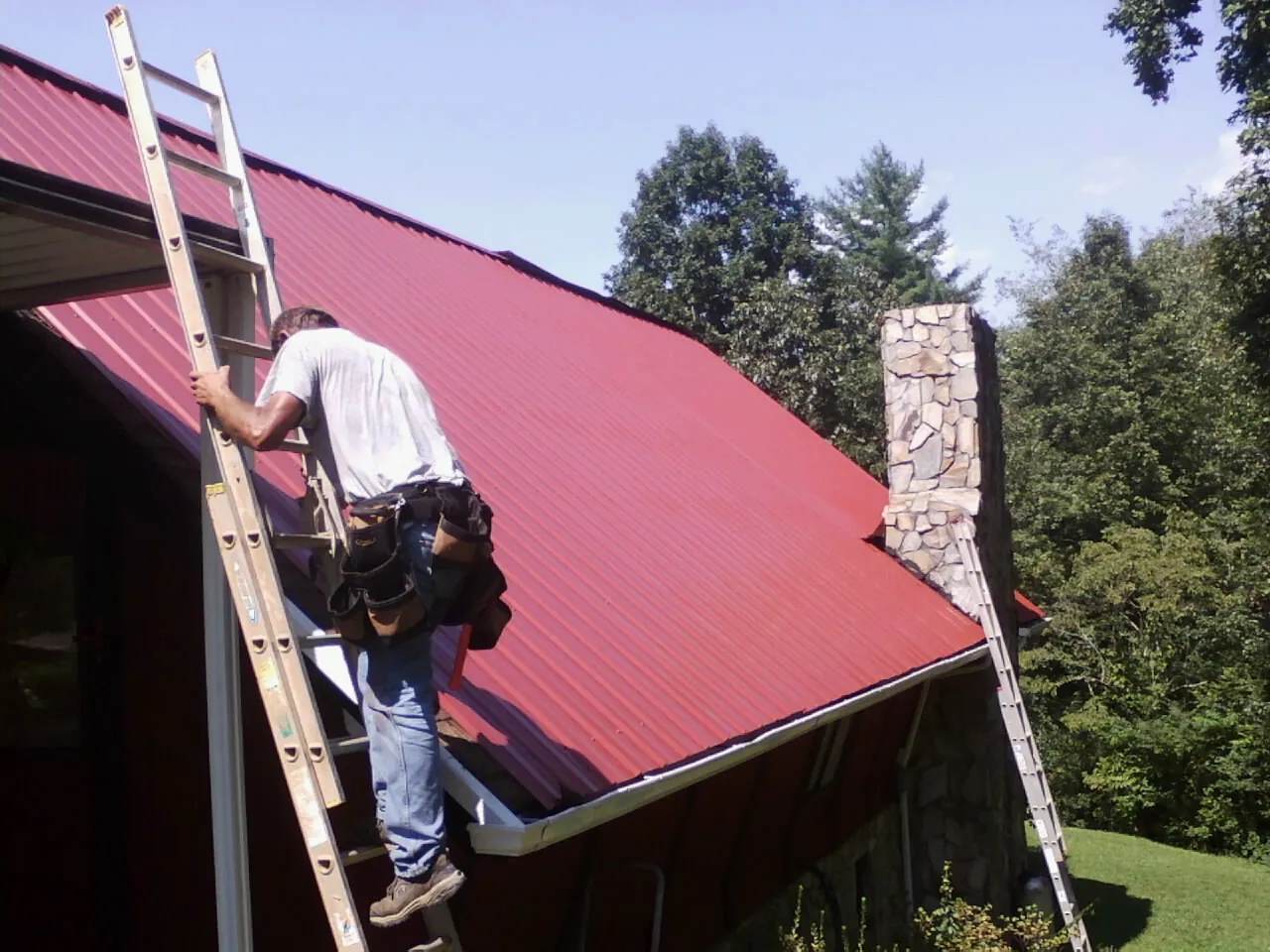 Roofer on a ladder beside a red metal roof and stone chimney — Soffit & Fascia Repair by Roofing Brothers in Bayonne